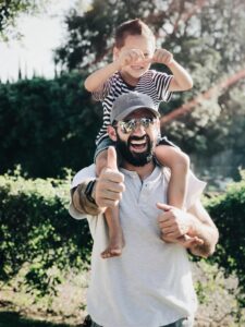 Man giving a thumbs-up with a child on his shoulders outdoors.